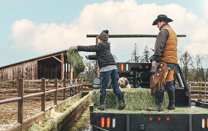 Image features a man, child, and dog standing on the back of a truck moving hay while wearing BOGS seamless boots.