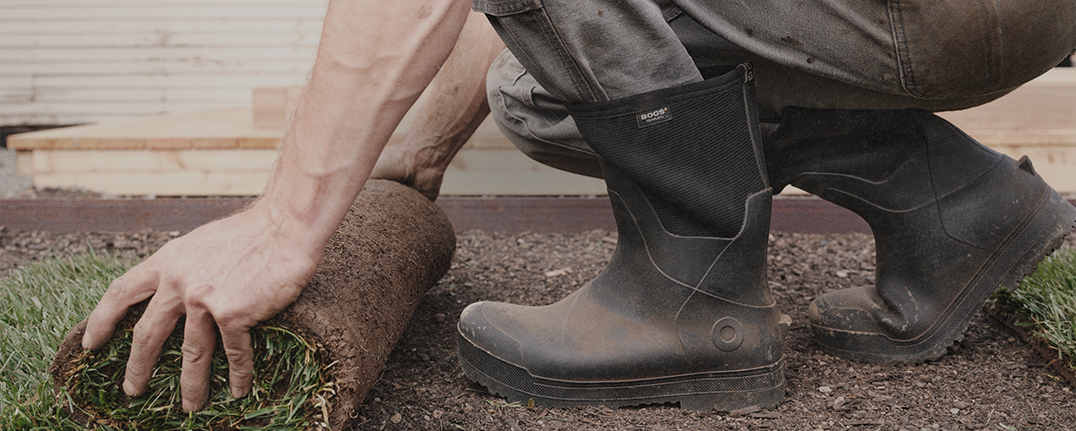 Image features a man in black boots rolling out sod. 