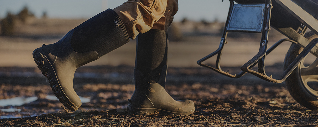 Image features a man walking in a muddly field wearing tall black boots. 
