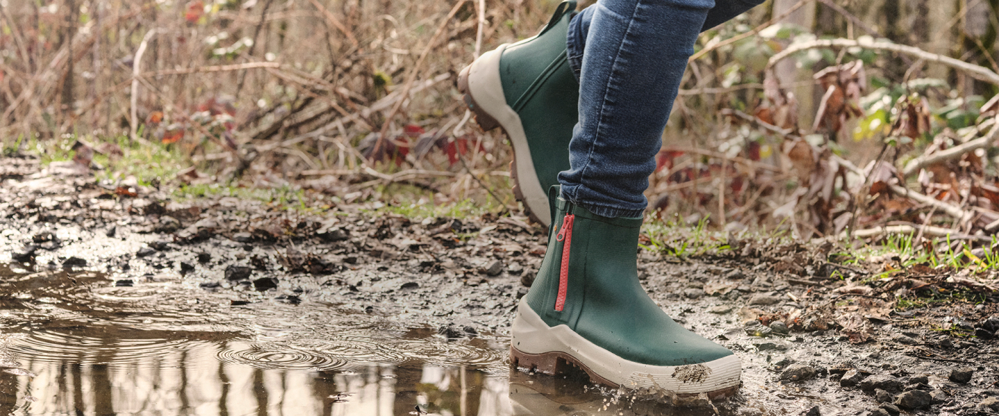 Image features a man in the woods bent down at a campfire wearing BOGS boots.