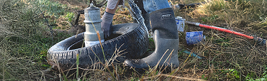Image features a medium-wide shot of a model working outside in front                  of a tire while wearing BOGS Seamless Construction boots.