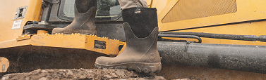 Image shows a close-up of a model stepping              up onto a construction vehicle while wearing muddy BOGS Seamless Construction boots.