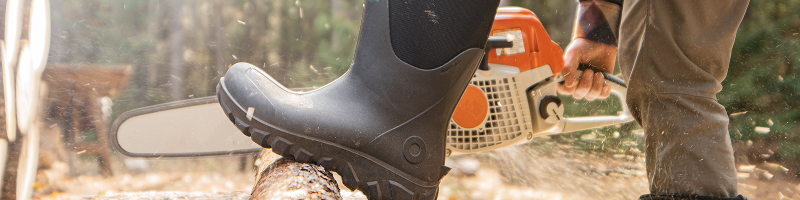 Image shows a close up of a BOGS boot on a model                  with their leg up on a log they are cutting with a chainsaw.