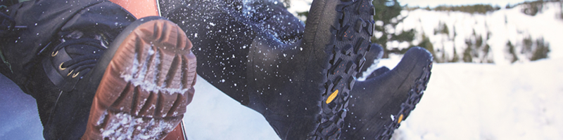 Image features a close-up of the bottom of BOGS snow boots                worn by two models sitting in the snow.