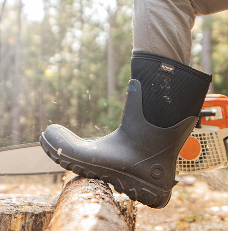 Image shows a close up of a BOGS boot on a model                  with their leg up on a log they are cutting with a chainsaw.